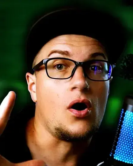 Reviewer in a black cap and glasses raising one hand toward the lens, mouth open, green backdrop