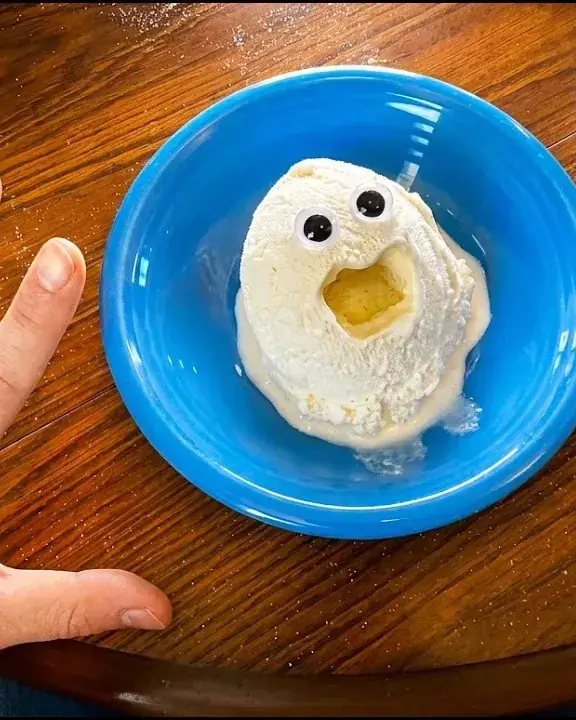 Overhead shot of a vanilla scoop with googly eyes and an open mouth on a blue plate, on a wooden table